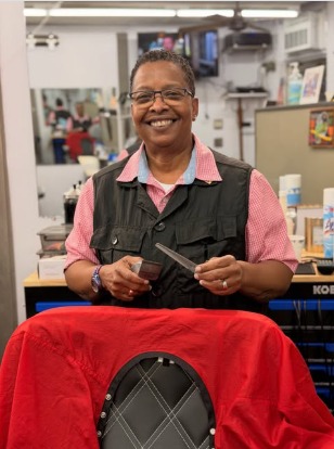 Mirna's Precise Edge holding a comb and shears beside a chair with a red cape